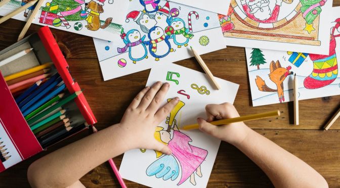 A view from above, looking down on a child's hands holding a yellow pencil and coloring in christmas scenes.